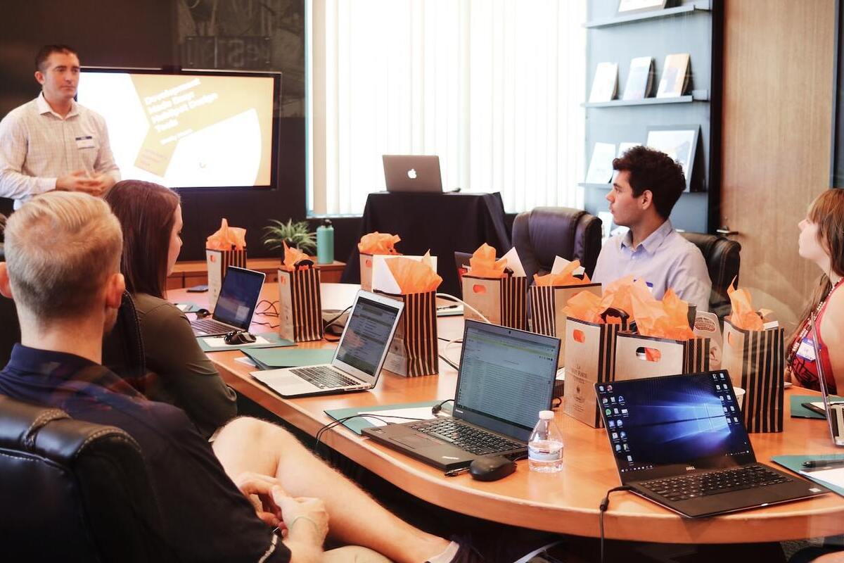 Reunião de trabalho: ambientes de marketing Equipe reunida em sala de reunião com líder em pé diante deles ao lado de tela com dados da palestra. Homens e mulheres com seus notebooks ao redor da mesa de madeira