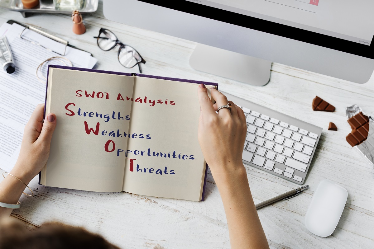 Mulher segurando livro: Análise SWOT para e-commerce Imagem de uma pessoa em frente ao computador segurando um livros com páginas brancas e escrito com SWOT Analysis.
