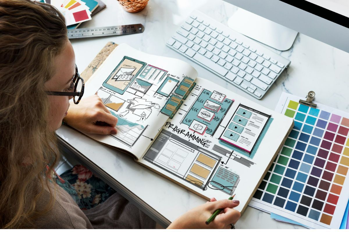 Mulher fotografada de cima com óculos de armação preta folheando apostila sobre marketing diante de seu computador em mesa branca de escritório com alguns objetos como uma régua de metal à sua esquerda e folha com paleta de cores impressa à sua direita