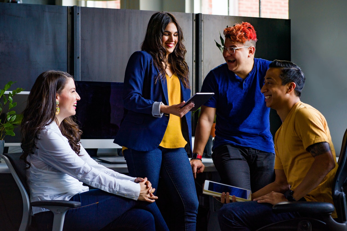 Quatro pessoas conversando: Como transformar clientes em promotores Na imagem mostra quatro pessoas sorridentes, onde duas estão com tablets nas mãos falando de negócios.