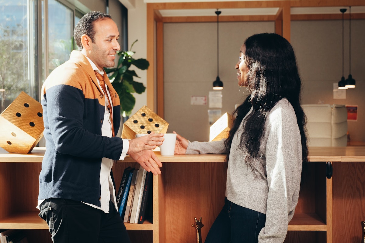 Duas pessoas conversando: Como transformar clientes em promotores A imagem apresenta uma mulher e um homem conversando, ao fundo tem uma estante com alguns objetos de decoração e livros.