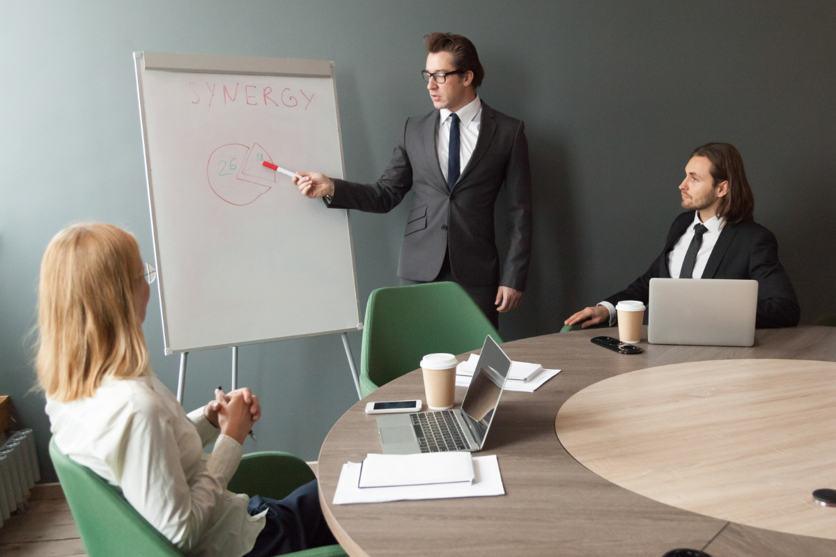 Reunião de trabalho: equipe de vendas Reunião de trabalho de equipe de vendas com homem e mulher sentados em cadeiras ao redor de mesa redonda e líder de pé apontando para painel com dados sobre sinergia