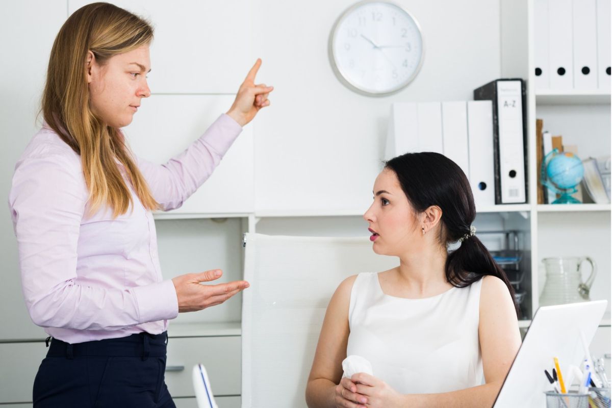 Duas mulheres em ambiente análogo a escritório onde uma está sentada e a outra de pé enquanto aponta para relógio em situação que lembra diálogo comum