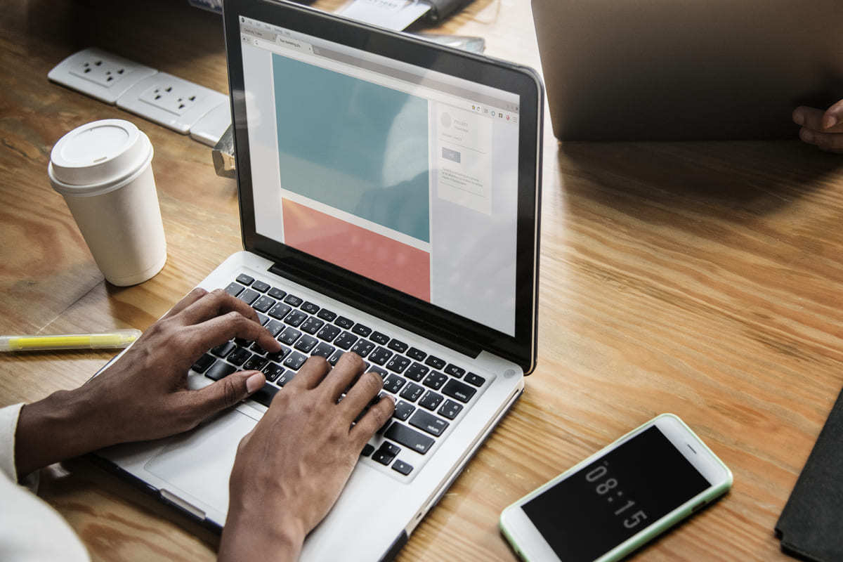 Pessoa usando notebook sobre mesa de trabalho em madeira com copo de café à esquerda e celular à direita