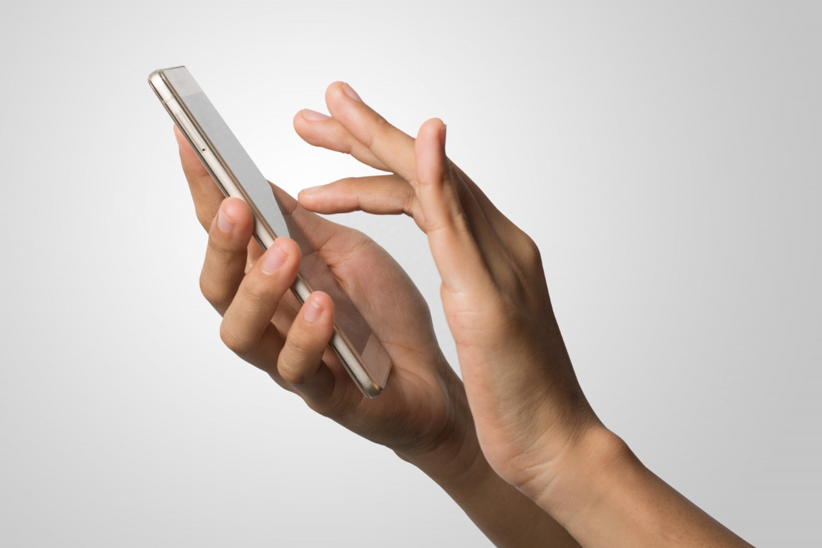 Mãos de pessoa segurando smartphone como se clicasse em algo na tela diante de fundo de imagem na cor branca e bordas em cinza