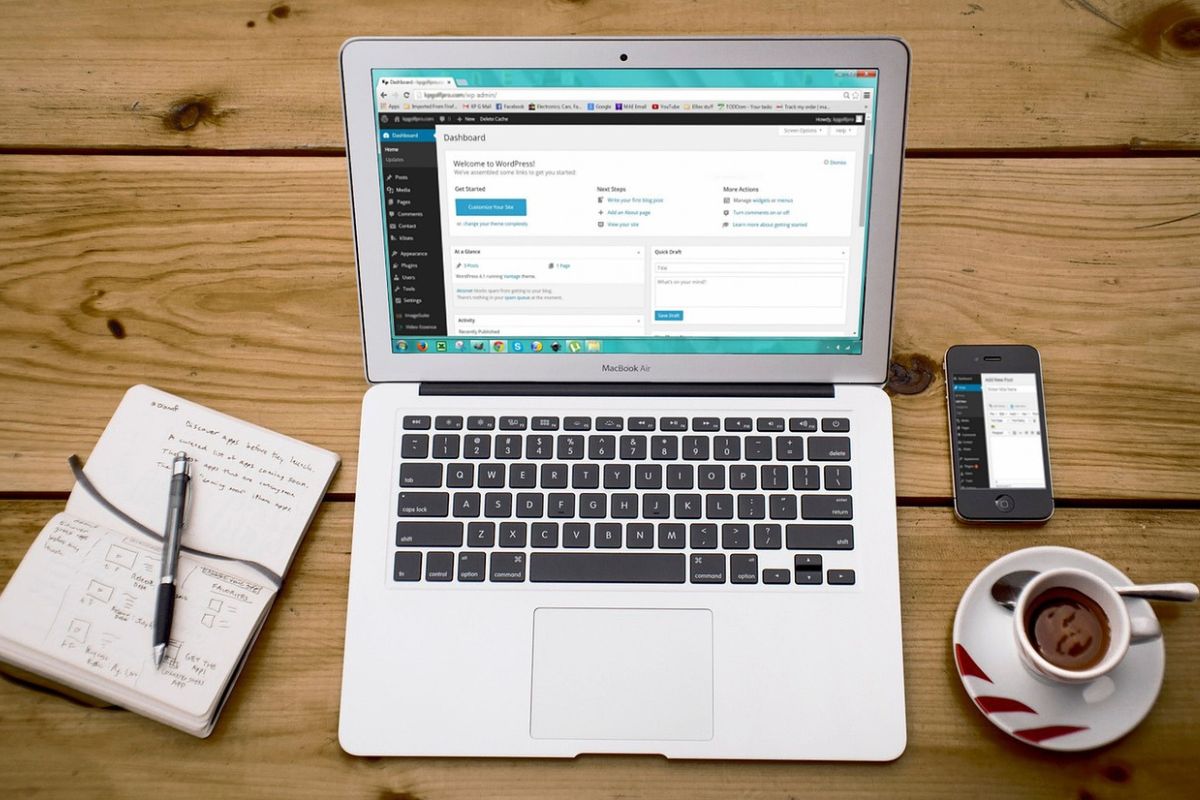 Notebook aberto: KPIs de SEO Na imagem está uma mesa de madeira, com um notebook aberto e na tela apresentando os dados do WordPress. Além disso, na mesma está um aparelho smartphone, uma xícara de café e também um caderno com anotações e uma caneta.