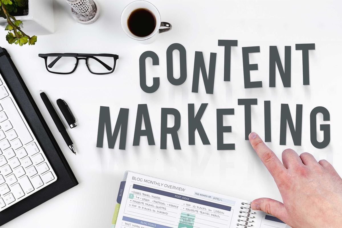 Homem apontando para algumas palavras: KPIs de SEO A imagem apresenta uma mesa branca de fundo, onde há um teclado, óculos, canela, planta, xícara de café e um caderno. Na mesma imagem está uma mão de homem apontado para as seguintes palavras Content Marketing.