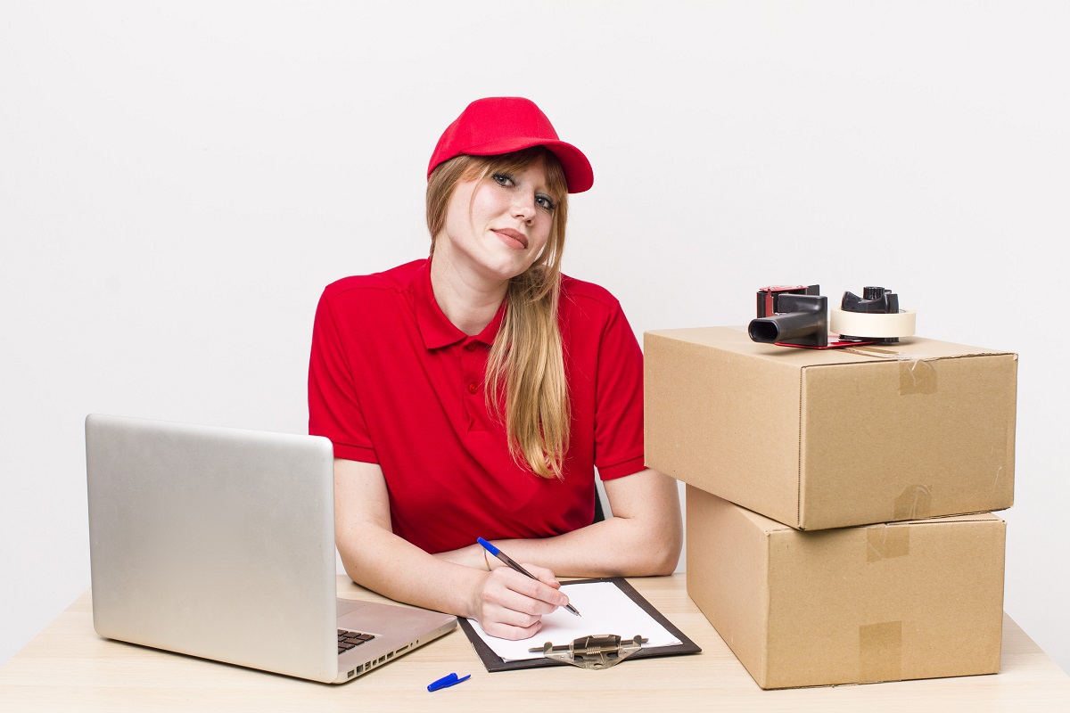 Mulher trabalhando: marketing para delivery Mulher com uniforme vermelho com boné vermelho usando prancheta ao lado de notebook e caixas ao lado que serão despachadas