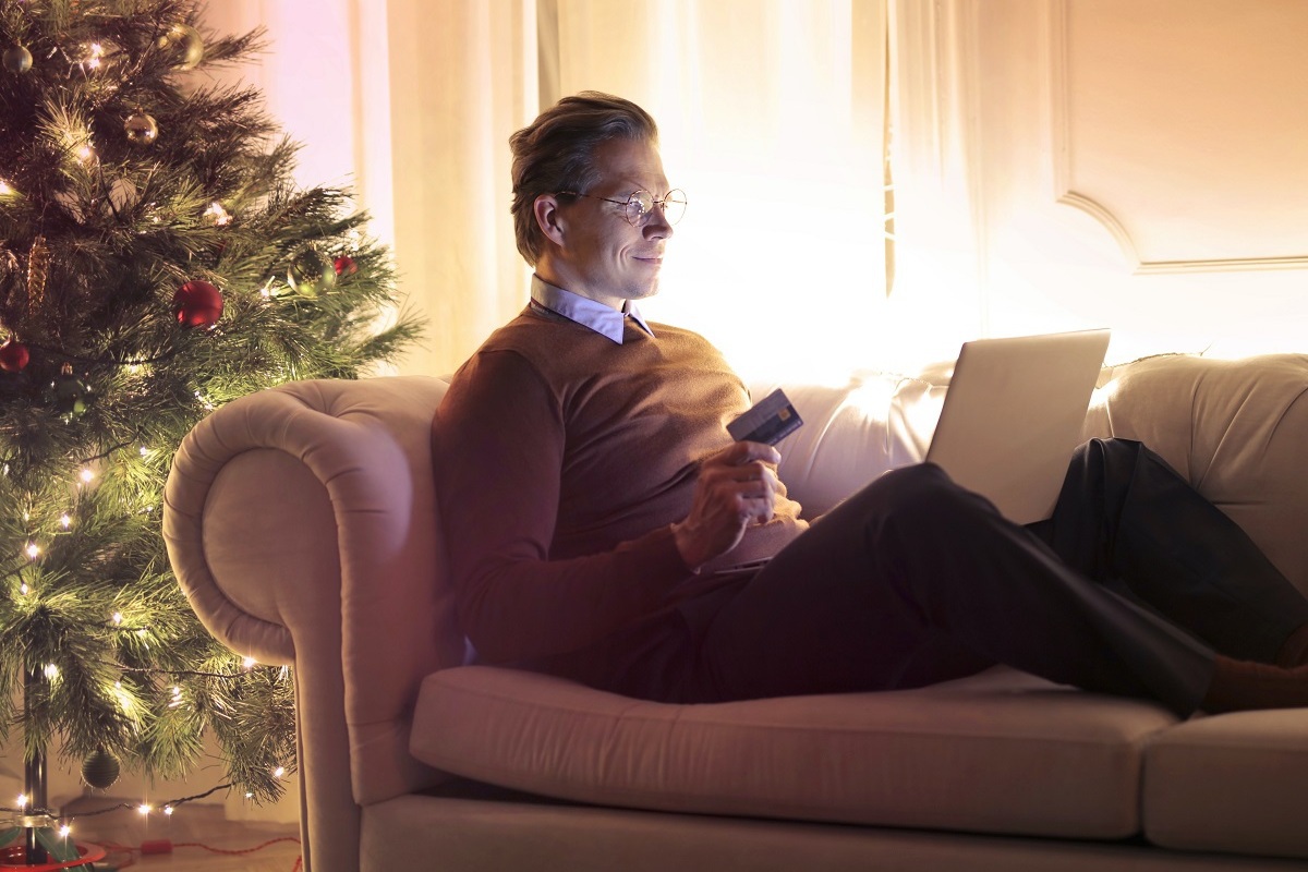 Compra online: melhore a conversão Homem sentado com pernas esticadas em sofá ao lado de árvore de natal com notebook no colo e cartão de crédito em mãos para fazer compra online