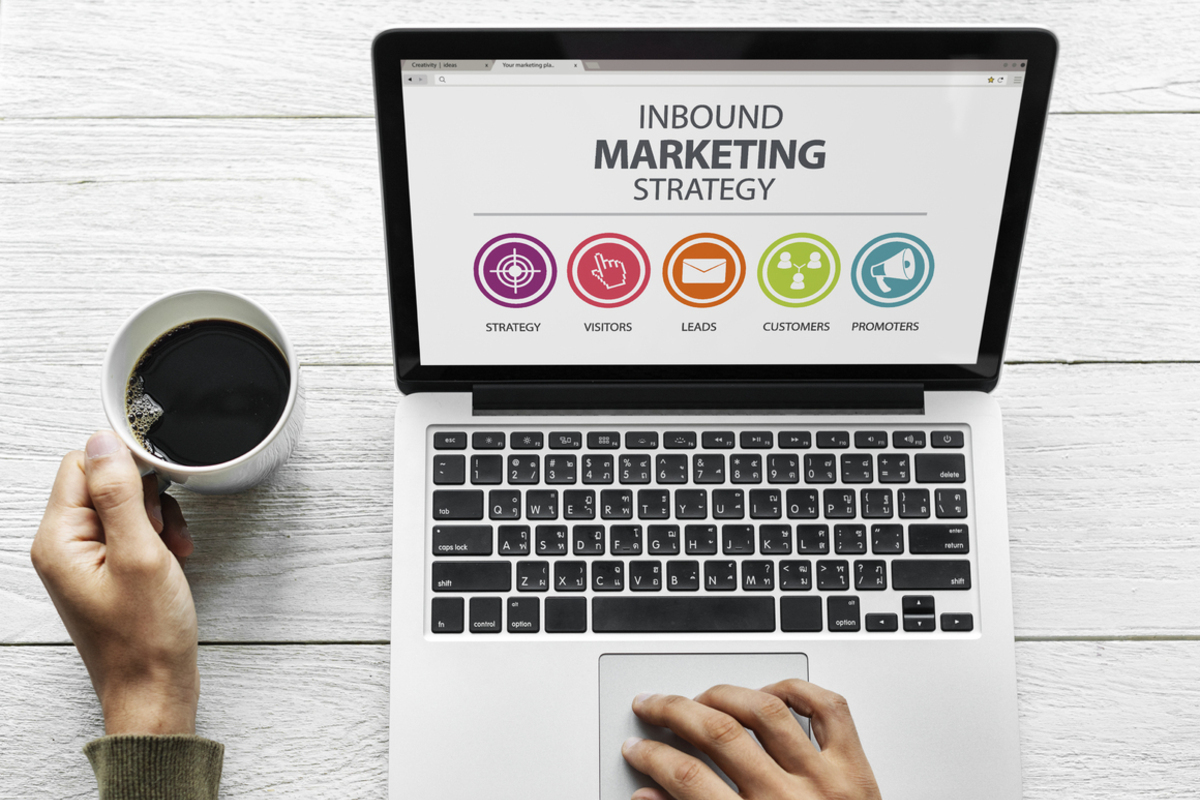 Pessoa trabalhando em notebook com mão direita e na mão esquerda uma caneca de café. Na tela do computador está aberta uma imagem que fala sobre Inbound Marketing e a mesa que segura os objetos é de madeira em cor clara, quase branca