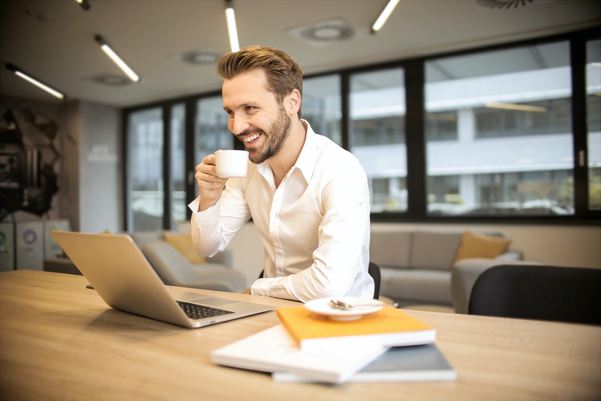 Homem segurando xícara de café: Social Styles Homem segurando uma xícara branca, encostado em uma mesa de madeira, onde se econtra livros, prato de sobremesa e um notebook prata.