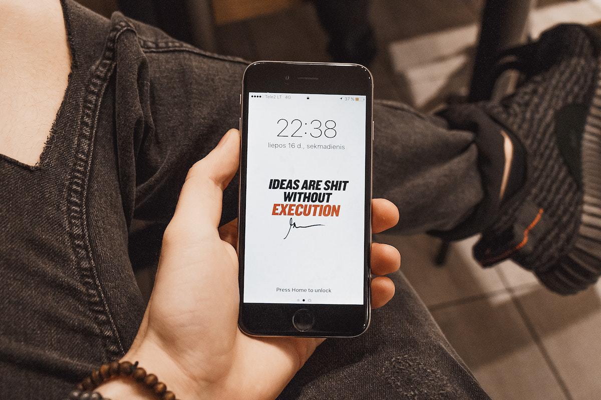 Pessoa segurando celular: texto publicitário Visão em primeira pessoa de de indivíduo segurando celular em tela de bloqueio, onde exibe horário 22:38h e frase em inglês "Ideas are shit without execution".