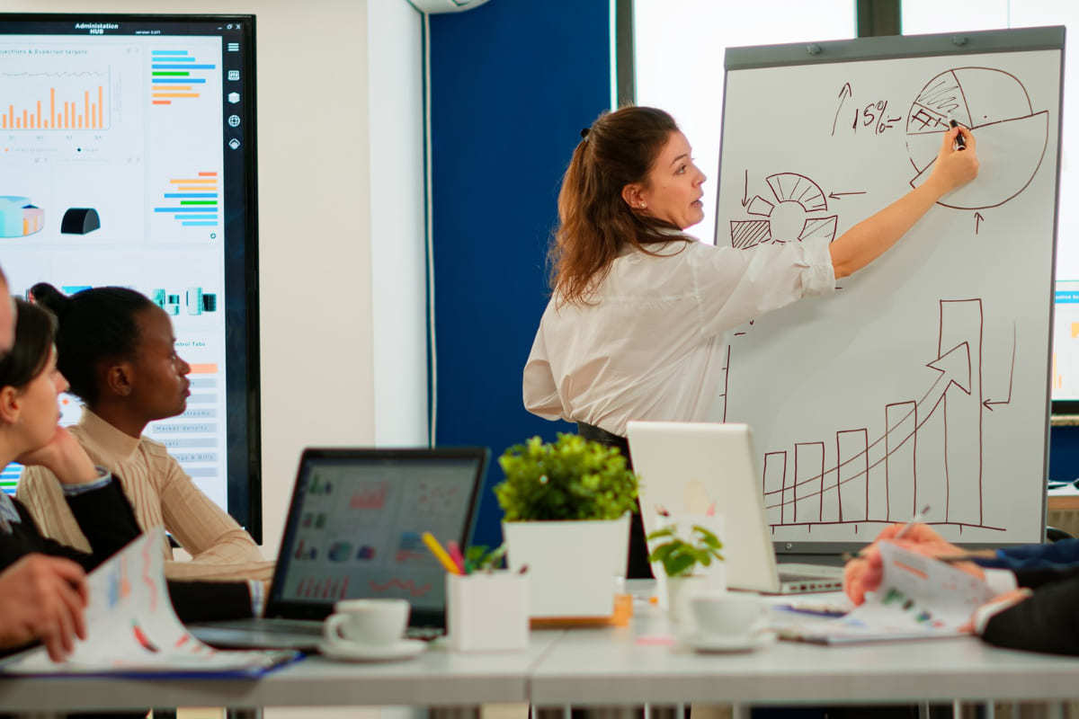 pessoas em reunião: canais de marketing mulher de pé, escrevendo em um quadro branco, enquanto seus colegas estão trabalhando sentados em uma mesa de reunião