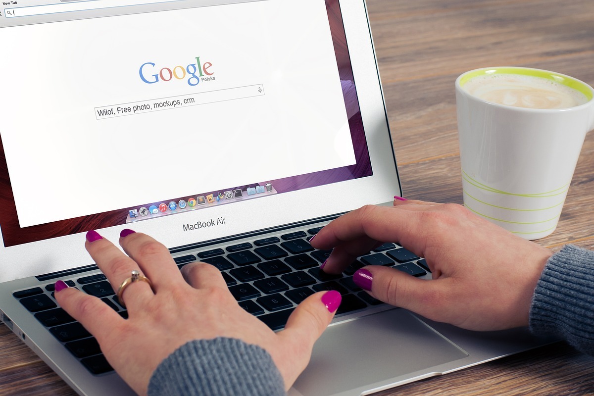 Mulher usando MacBook: criar conta Google Mulher usando MacBook Air sobre mesa de madeira e com copo de café ao lado