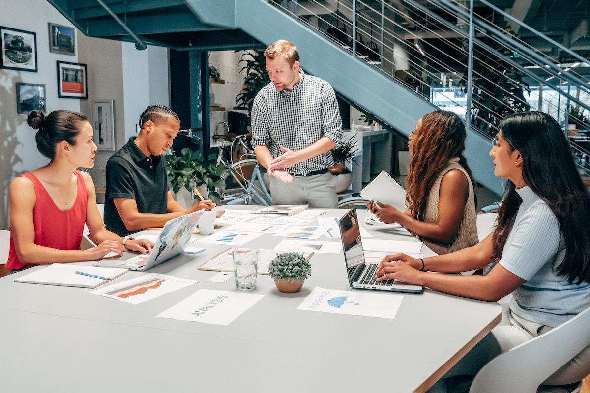 Time: 4 As do marketing Três mulheres e um homem sentados ao redor de mesa grande em local de trabalho usando notebooks e com vários gráficos impressos, calculadoras e canetas espalhados ao redor de todos com líder diante de todos falando e gesticulando sobre tema