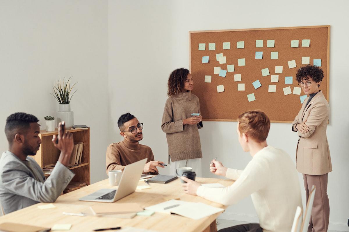 Três homens sentados ao redor de mesa retangular de madeira em reunião de trabalho com duas mulheres de pé liderando o evento diante de quadro com vários post-its colados no mesmo