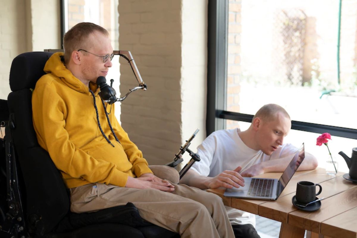 Homens com notebook: acessibilidade digital Homem em cadeira de rodas repleta de recursos com outro homem abaixado na sua frente verificando algo em notebook que está em cima de mesa de centro de madeira