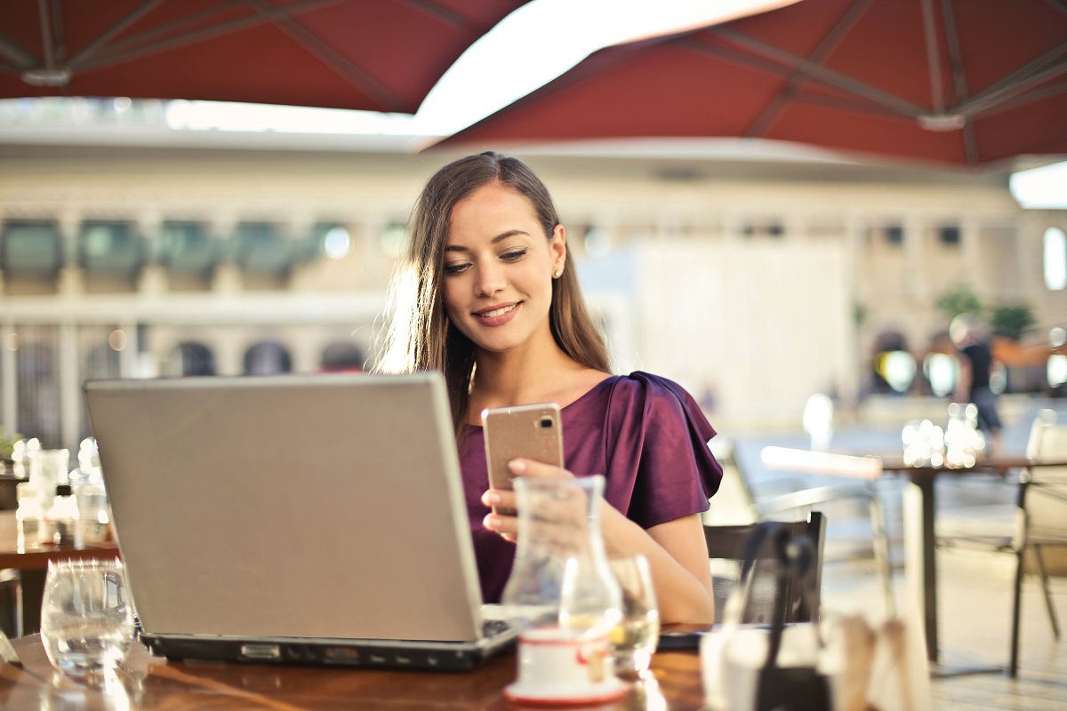 Mulher em área de piscina de hotel de luxo segurando smartphone em uma das mãos e com notebook sobre mesa de madeira com jarra de vidro com suco e copo de água ao seu redor