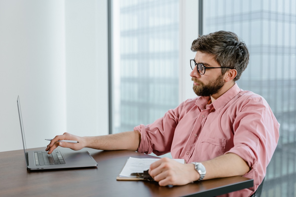 Homem de camisa social cor-de-rosa sentado diante de mesa de madeira usando notebook e segurando prancheta com folhas a4, na frente de janela de vidro grande voltada à outro edifício