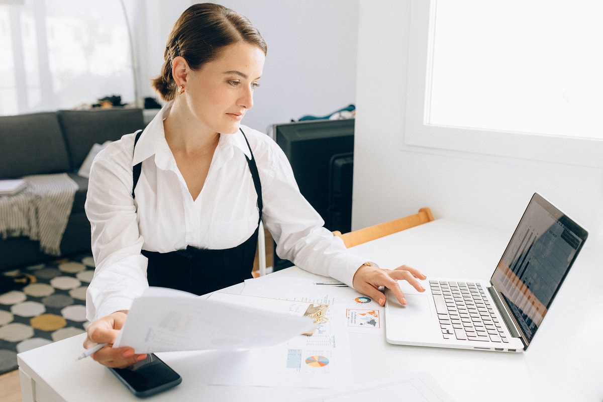 Mulher em home office usando notebook sobre mesa e segurando folhas a4 com móveis de sala de estar ao fundo da fotografia em desfoco
