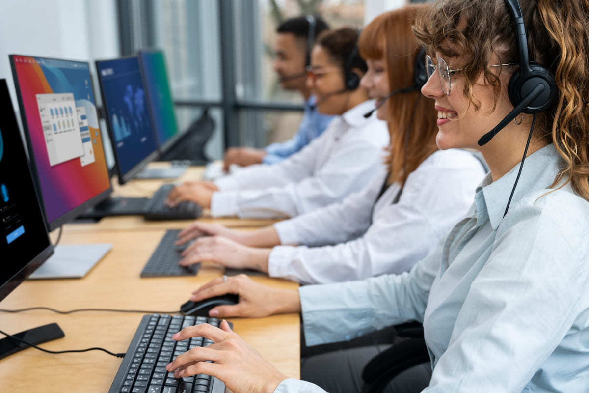 Local de trabalho: atender clientes mais rápido Três mulheres e um homem sentados diante de suas mesas lado a lado e usando computadores e fones de ouvido para atender clientes mais rápido