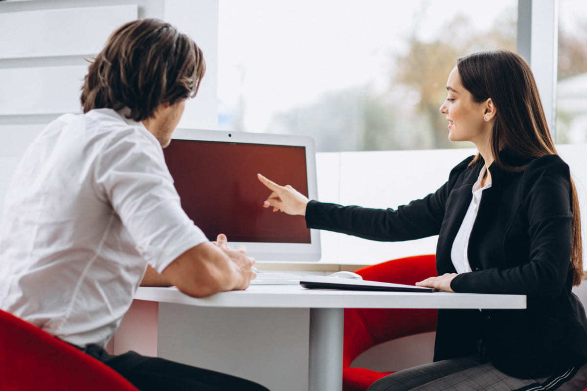 Homem e mulher sentados frente a frente em pequena mesa branca discutindo sobre algo em computador de mesma cor sobre a mesa em escritório