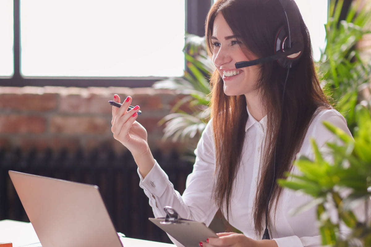 Mulher sorridente: atender clientes mais rápido Mulher sentada diante de mesa com notebook e fone de ouvido sorridente enquanto atende cliente