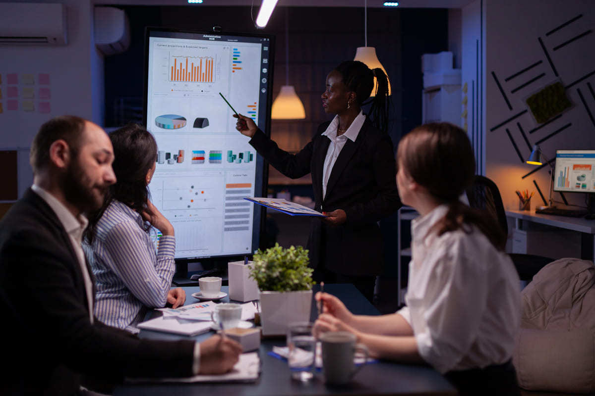 Mulher de pé em reunião de trabalho exibindo gráficos em tela para colegas, que são duas mulheres e um homem, sentados à sua frente ao redor de mesa retangular preta com xícaras de café e gráficos impressos espalhados sobre a mesma