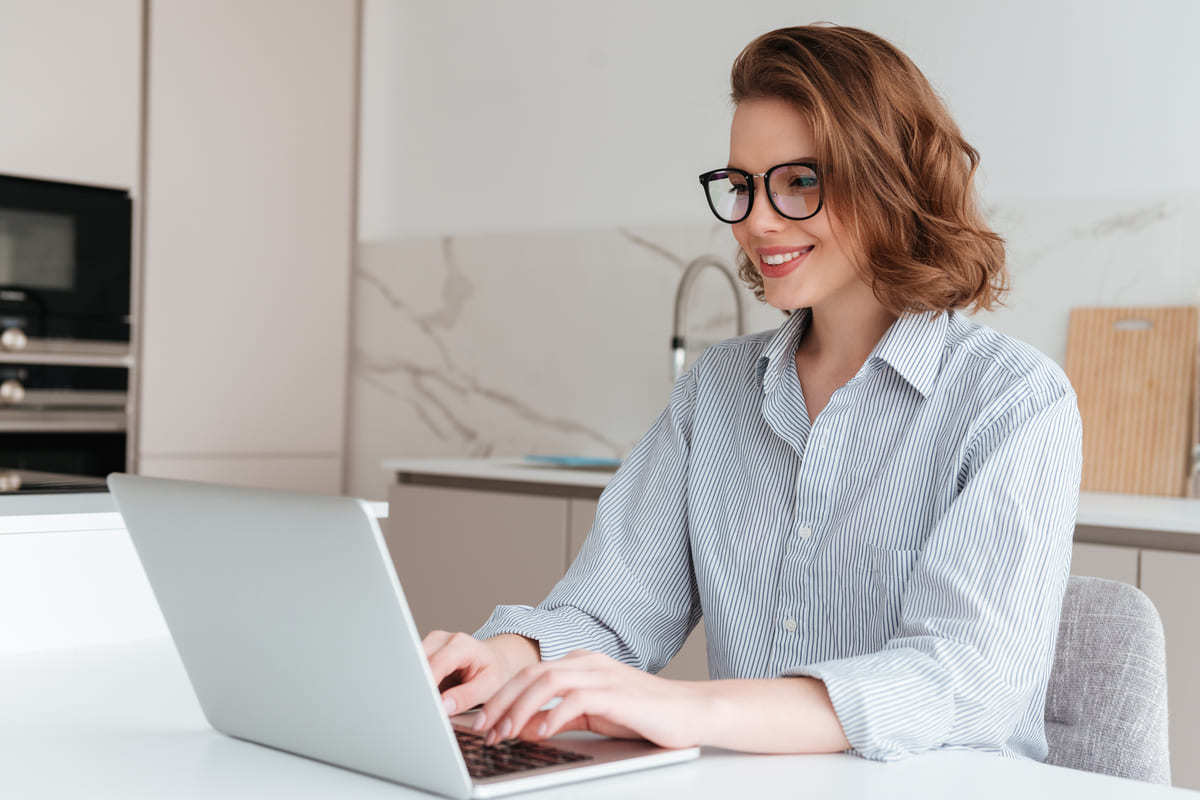 Mulher com camisa social clara de mangas compridas e botões sentada em cadeira de escritório diante de mesa branca usando notebook cinza na frente de armário com pia