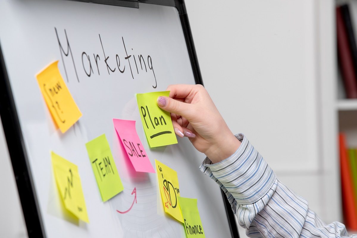 Pessoa colando post-it em painel branco com nome Marketing que tem outros adesivos colantes fixados ao mesmo