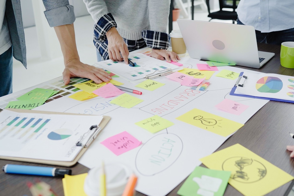 Gráficos: Brand content vs marketing de conteúdo Pessoas de pé na frente de mesa de mesa de madeira com várias folhas e post-its espalhados sobre a mesma e gráficos em pranchetas