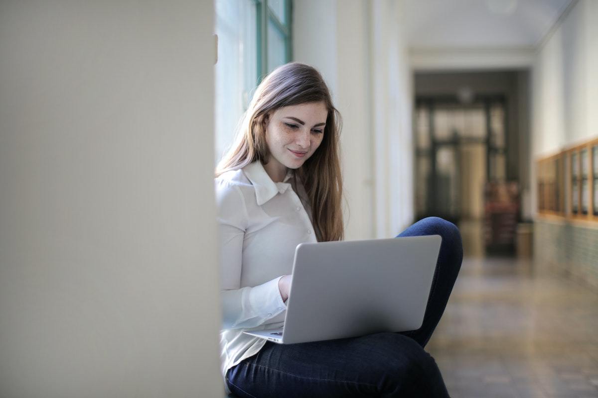 Mulher sorridente: Branding para IES Mulher sentada diante de grandes janelas de vidro em corredor que estão em desfoco no fundo da imagem segurando notebook cinza em seu colo com ar sorridente