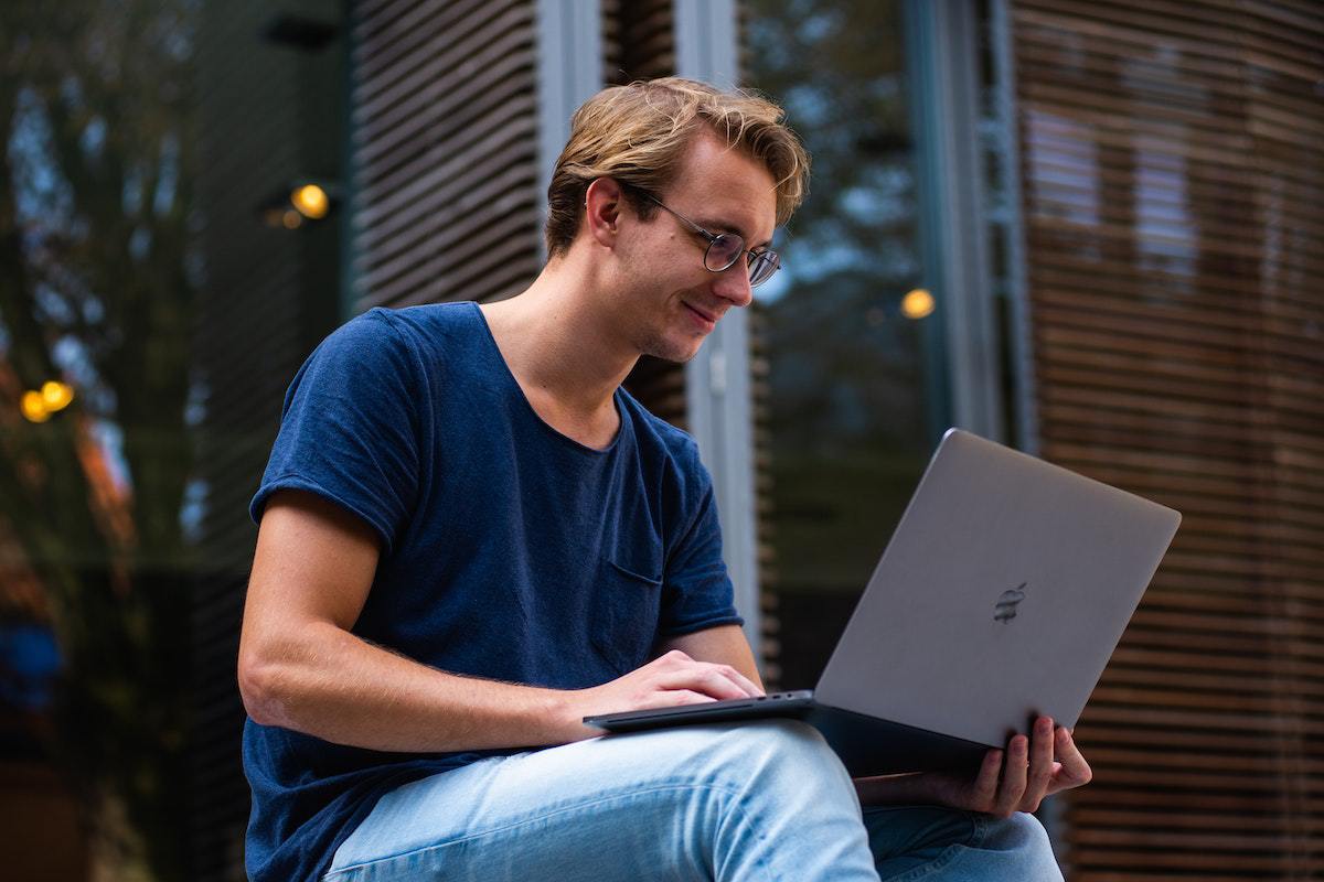 Homem sentado diante de grandes janelas de vidro em desfoco no fundo da imagem segurando notebook cinza em seu colo com ar sorridente