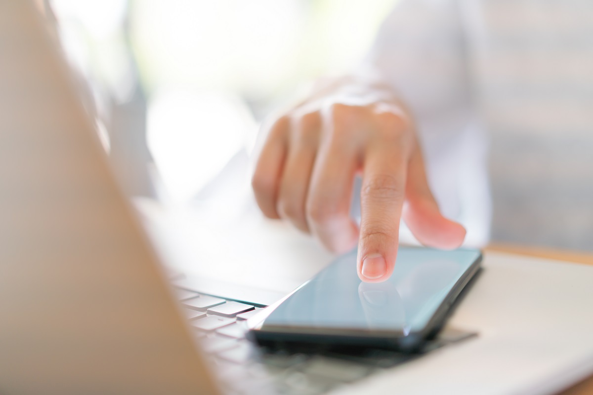 Mão feminina em foco na fotografia enquanto toca tela de celular que está sobre teclado de notebook cinza