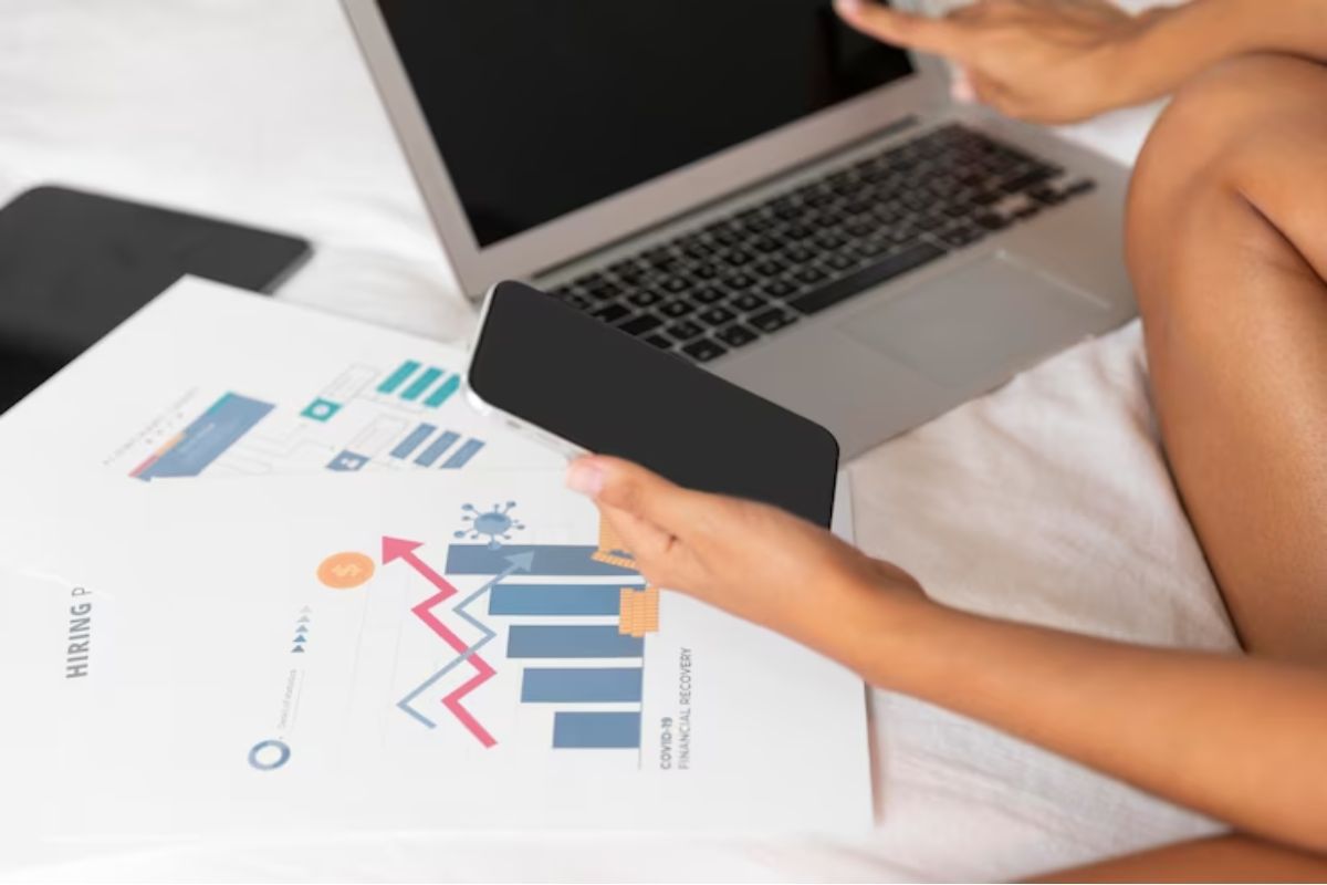 Aprenda a calcular metas de vendas e saiba o que levar em conta ao fazê-lo Mulher segurando celular e com mãos sobre gráficos impressos em folhas a4, além de notebook cinza à sua direita