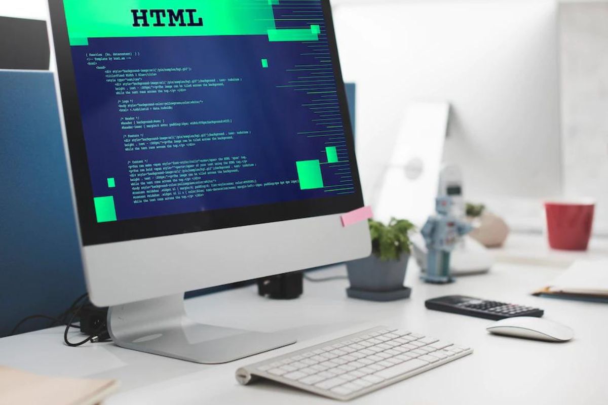 Computador branco em cima de mesa branca com código HTML em sua tela e objetos como canecas e celulares ao redor do teclado