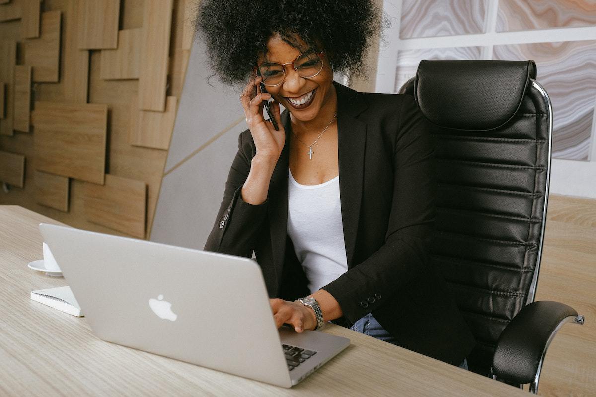 Mulher em escritório com roupa social sorri enquanto fala ao telefone e usa notebook