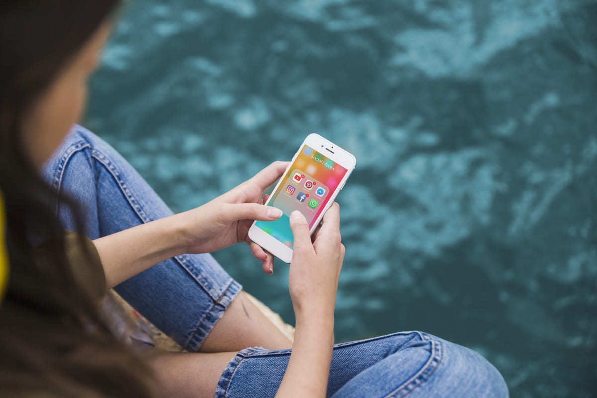 Mulher sentada de pernas cruzadas em banco de cimento e usando celular, que exibe menu de redes sociais na tela