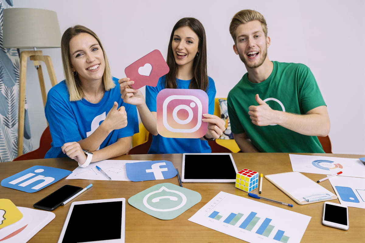 Duas mulheres e um homem sentados diante de mesa de madeira fazendo recortes em papéis com símbolos de redes sociais impressos enquanto sorriem