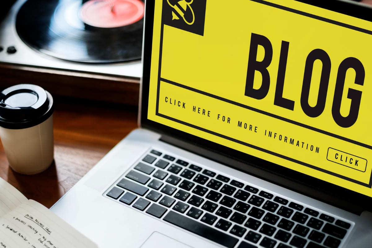 Notebook em cima de mesa com montagem com nome blog na tela ao lado de copo de café