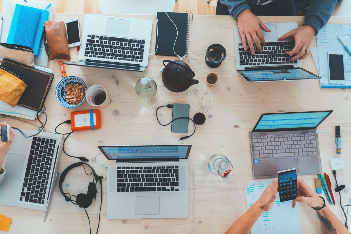 Dois homens, um diante do outro, usando notebook e smartphones com mais notebooks ao redor e canecas com café e bowls com grãos