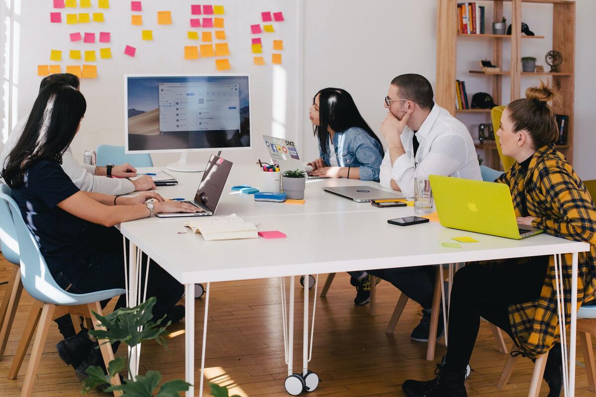 Homens e mulheres sentados ao redor de mesa branca em escritório olhando para TV com tela de notebook espelhada com alguns post-its acima da mesma