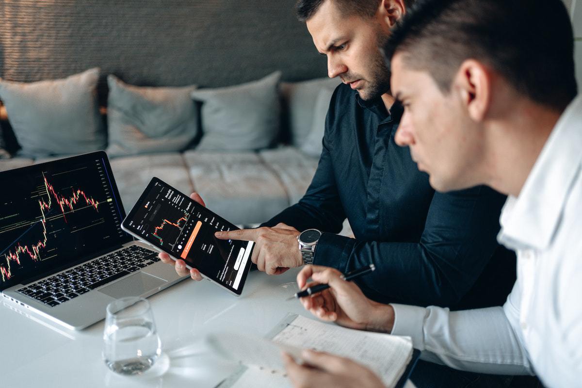 Dois homens sentados diante de mesa analisando gráficos em notebook e tablet