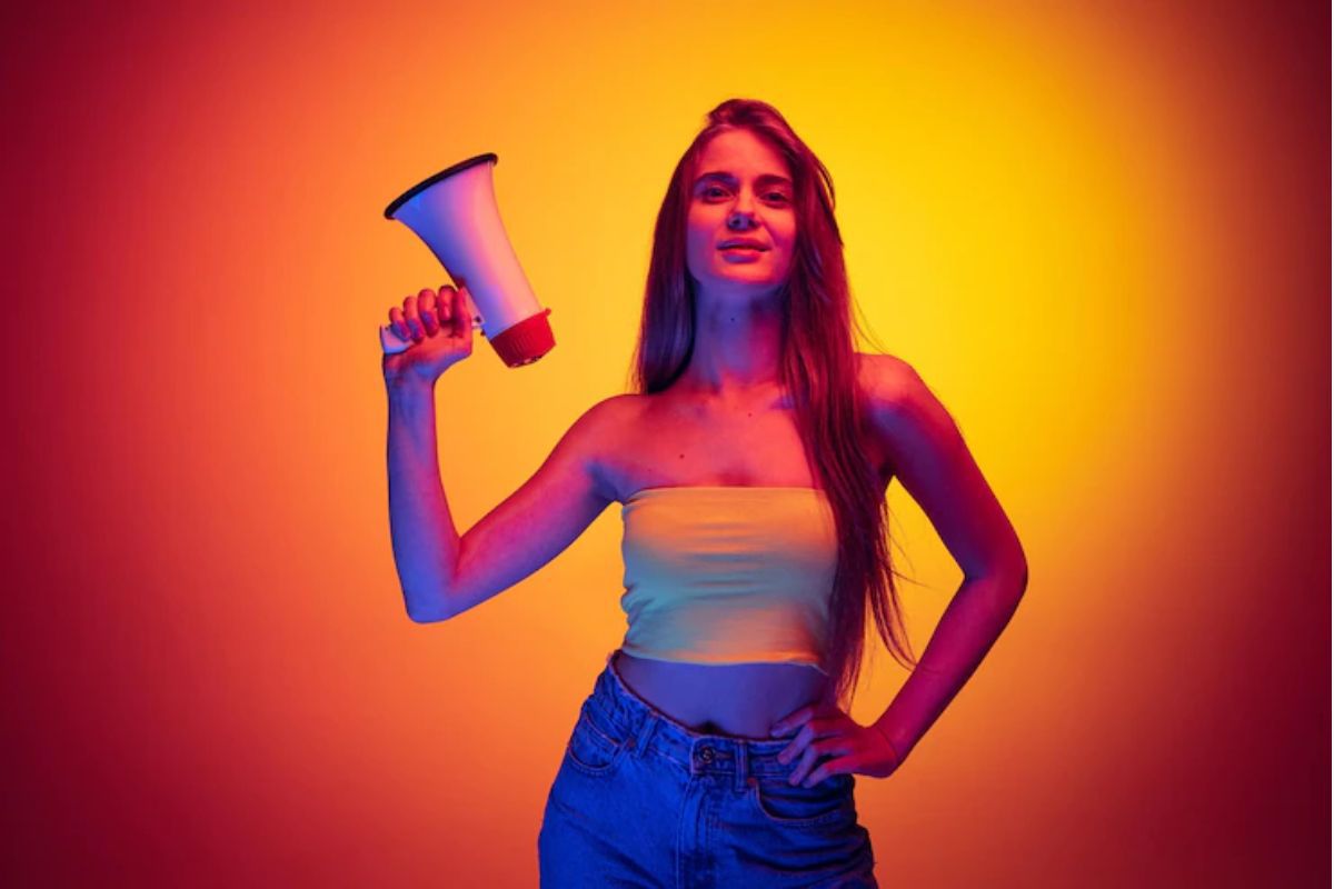 Mulher com megafone na mão esquerda e a outra mão na cintura em pose para foto diante de parede laranja com holofote no meio