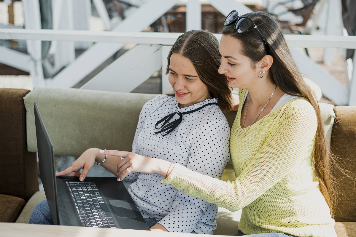 Duas mulheres com notebook: Conversão no e-commerce Duas mulheres sentadas em sofá de sala com notebook no colo de uma delas enquanto a outra aponta para a tela do mesmo conversando sobre algo