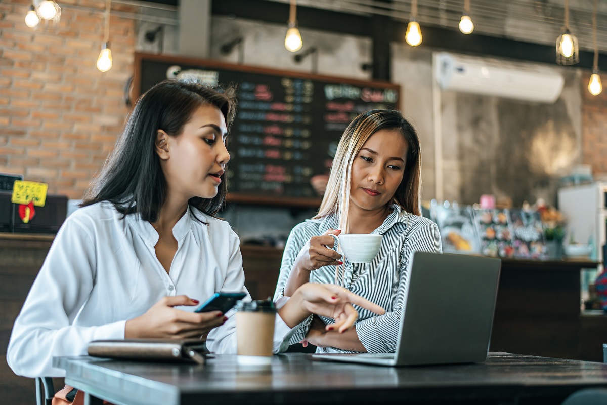Duas mulheres sentadas diante de mesa em interior de lanchonete apontando para tela de notebook e debatendo algo
