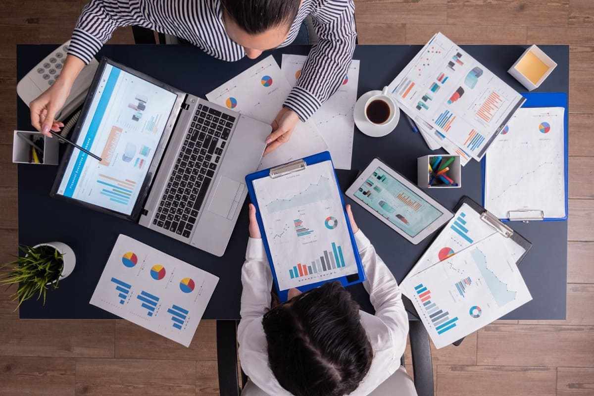 Escritório: criação publicitária Homem e mulher sentados em escritório, um diante do outro e um em cada lado da mesa analisando dados em notebook e em folhas impressas espalhadas na mesa