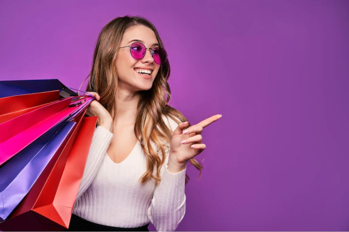 Mulher sorridente: cross sell Mulher sorridente segurando sacolas de compra diante de parede roxa com óculos de sol de lente também roxa e apontando para a sua esquerda