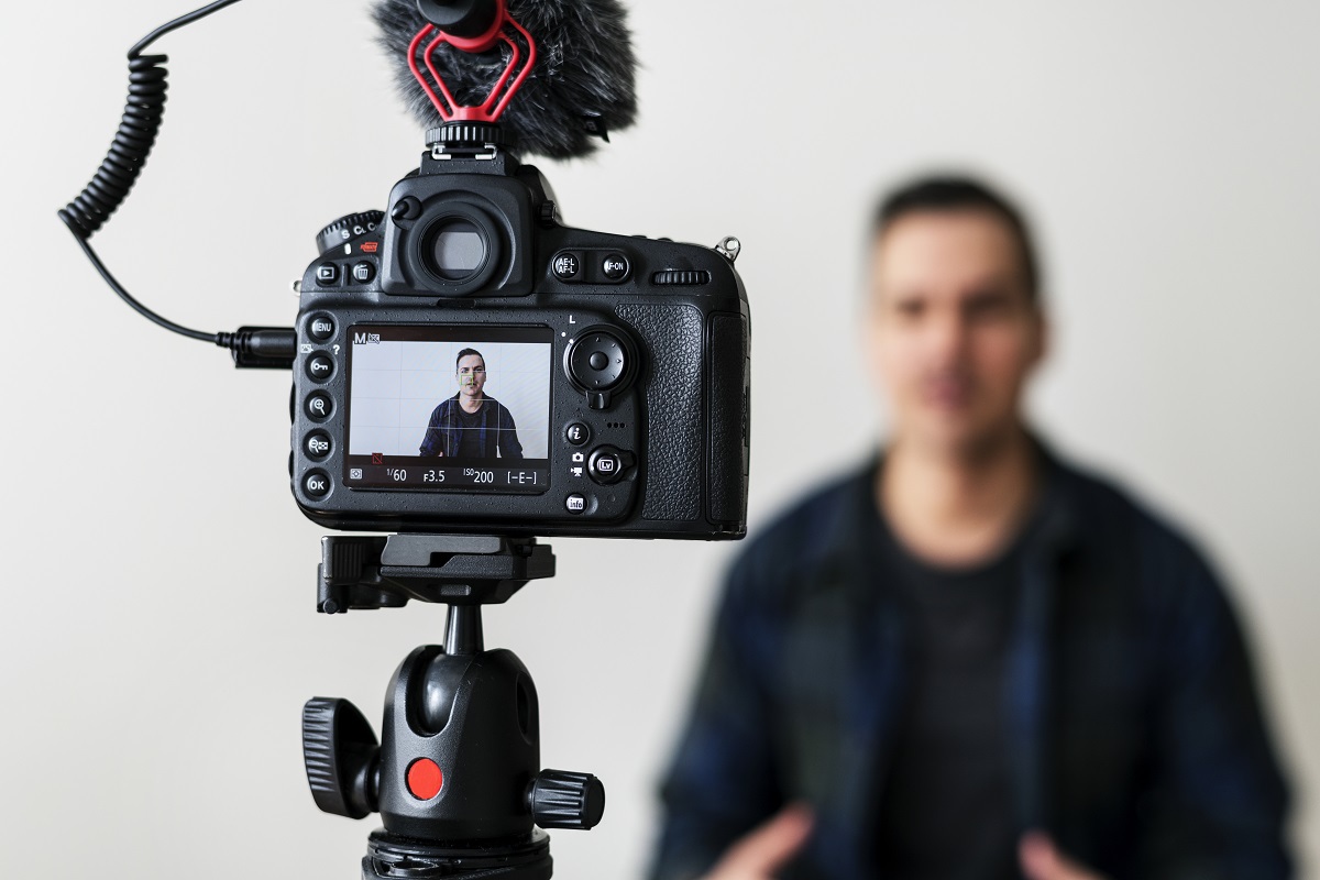 Homem de preto gravando vídeo diante de câmera semi profissional em pedestal com foco na pequena tela do aparelho