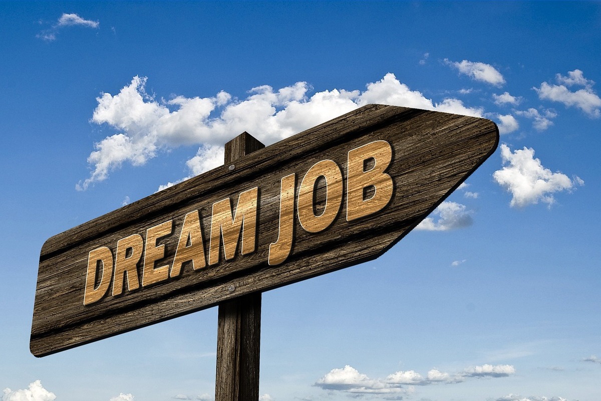 Trabalho dos sonhos: employer branding Placa de madeira em formato de seta apontando para a direita com a frase DREAM JOB escrita na mesma e ao fundo o céu azul com algumas nuvens dispersas